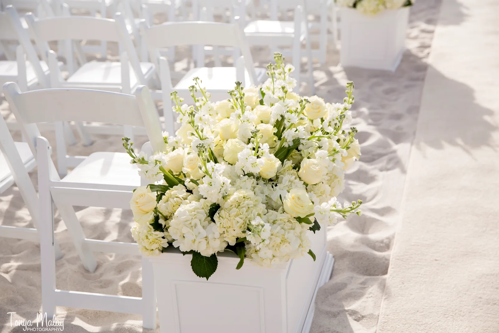White wedding aisle and floral pedestals leading to a draped ceremony structure on the beach
