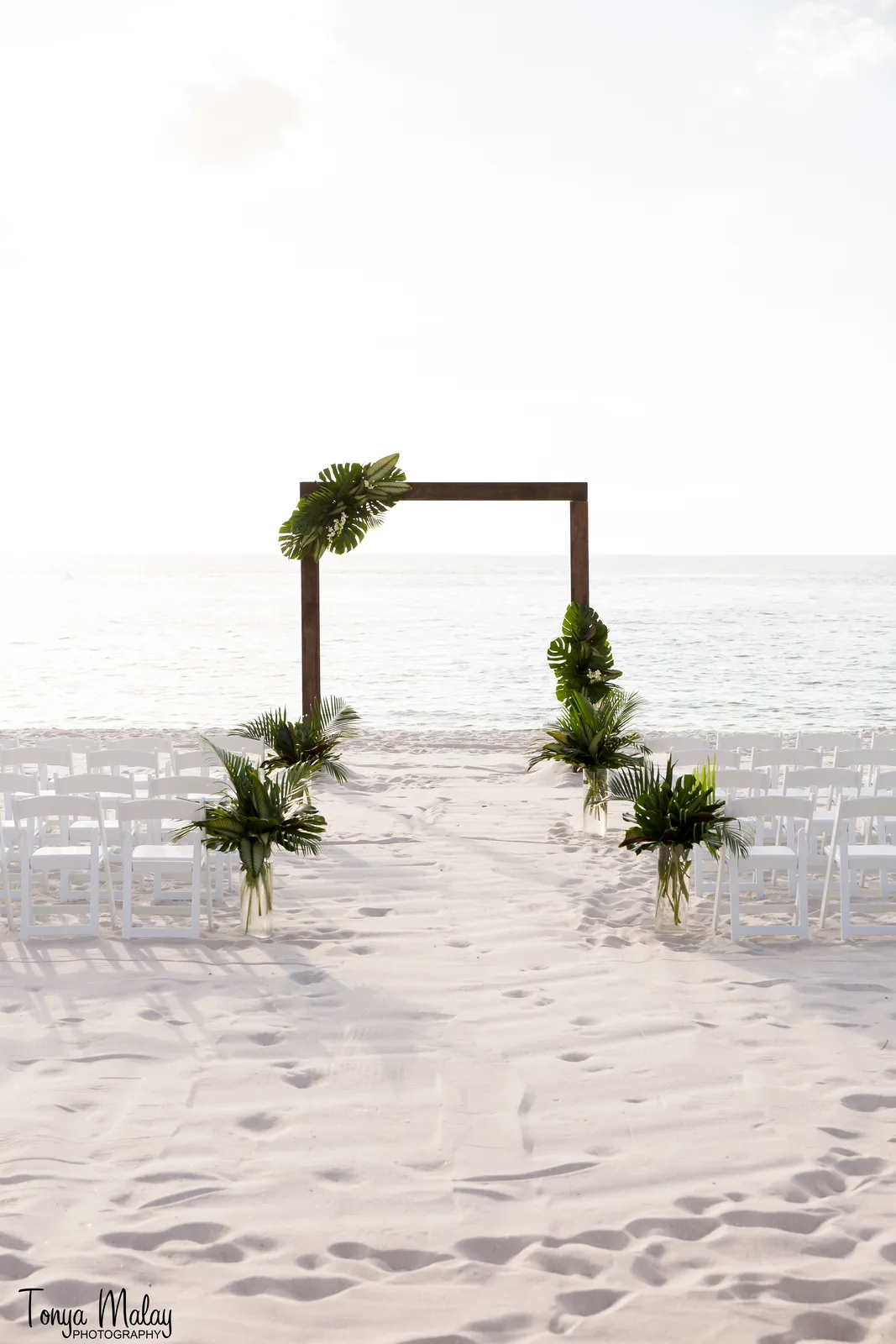 Minimalist wood ceremony arch with monstera and palm arrangements on white sand