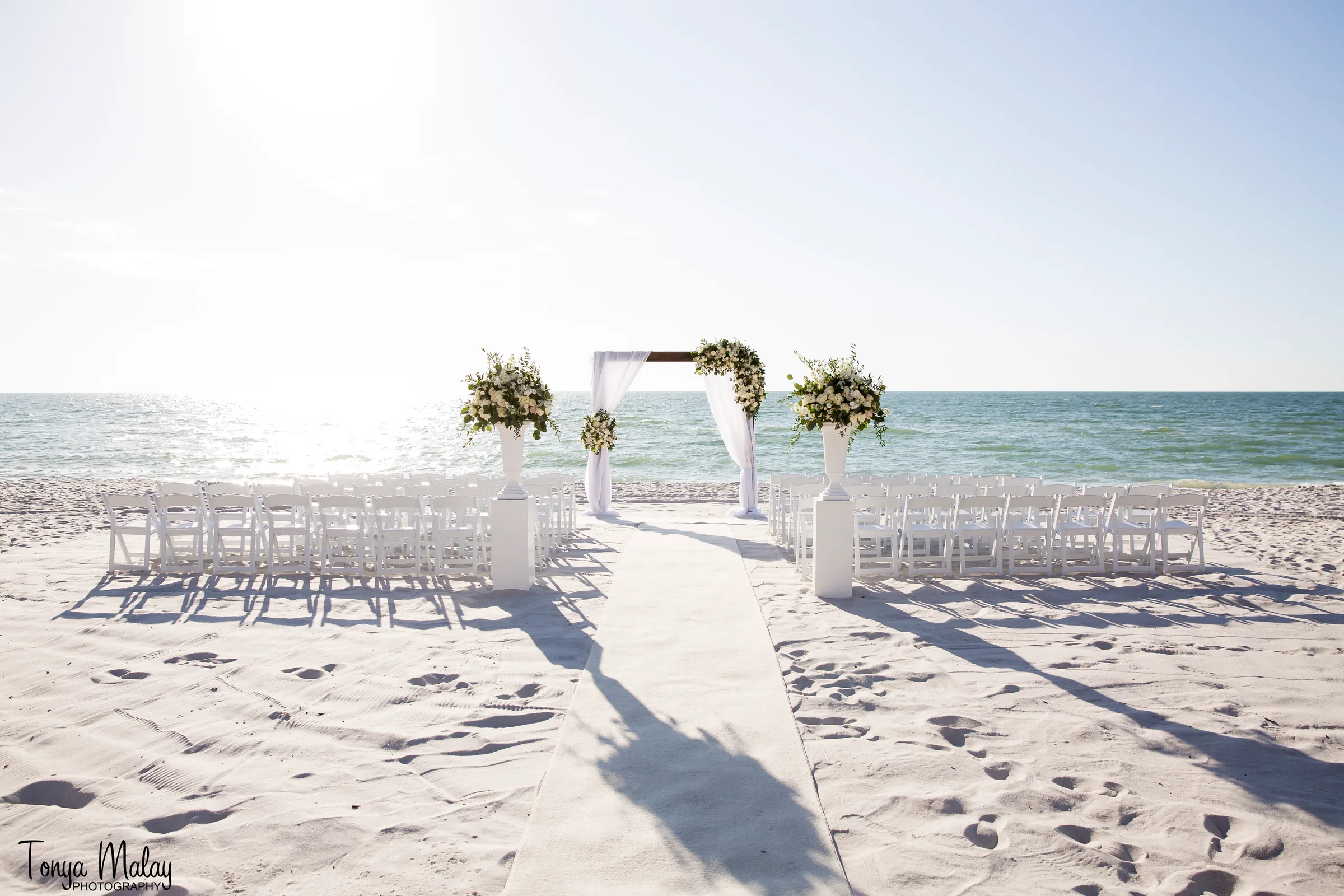Beachfront wedding ceremony with a white draped arch, urn florals, and a long aisle to the water