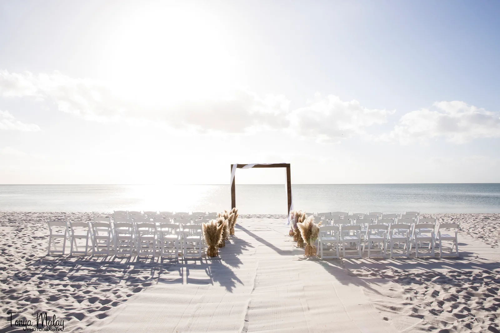 Sunlit beach ceremony with arch and pampas grass aisle arrangements